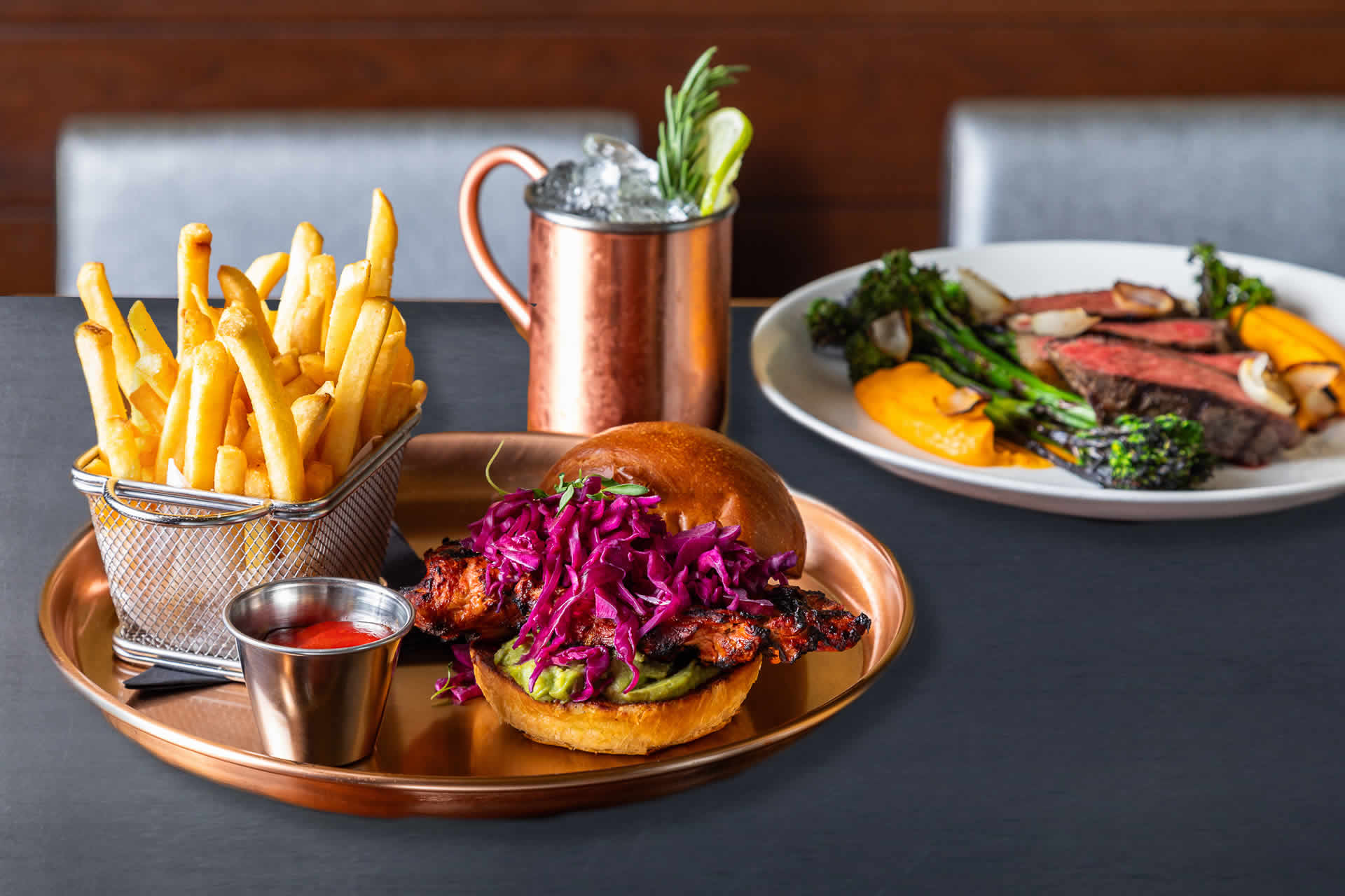 Chicken sandwich and french fries next to a plate of steak and broccoli with a Moscow mule.